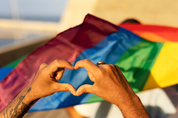 Hands making heart sign in front of rainbow flag.
