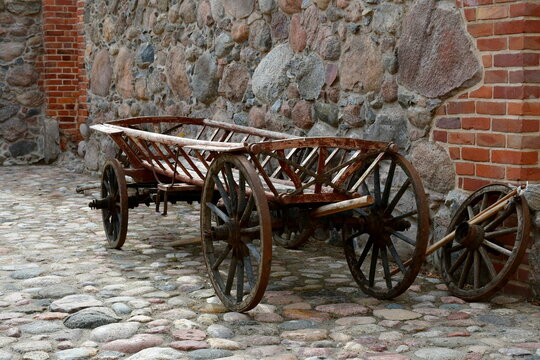 A close up on an old wooden cart made out of logs, planks, and boards and used to transport goods or crops seen next to a red brick wall and some stone fence on a sunny summer day in Poland