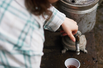 The lady turns on the tap and pours tea from an old samovar. Dirty wet floor. Unsanitary conditions on a camping trip. Traditions of tea drinking. View from above. Selective focus.