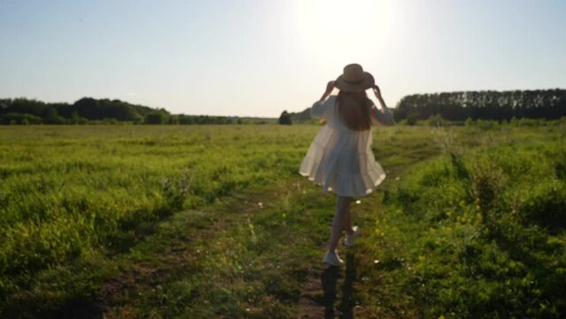 Back view of unrecognizable happy woman in straw hat and white dress joyfully running bouncing along in countryside field toward sun in summer evening during sunset. Shooting in slow motion.