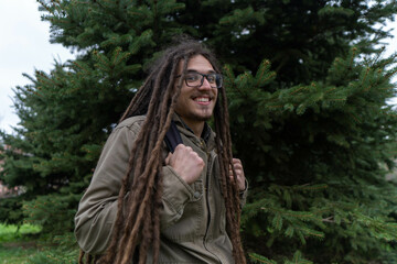 Young smiling boy with long dreadlocks posing next to trees. Traveler male wearing warm clothes posing in nature. Concept of scout, research, camping, travel and survival in nature.