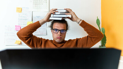 a young college student holding a pale of books on his head with a weary face, looking at laptop,...
