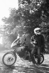 Fototapeta na wymiar rider guy use smart phone in jeans biker jacket and helmet sit on tourist touring motorcycle. outdoors, dual sport adventure concept, vertical photo, dirt road, black and white