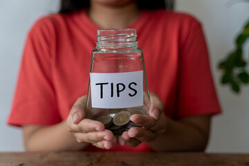 Glass jar with a coin in the woman's hand receives tips from the service in the work done. The...