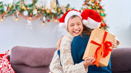 happy and loving mother and son, dressed in a Santa Claus hat, hug at home, sitting on the sofa against the background of a Christmas tree. child and woman are laughing happily while holding gift box.