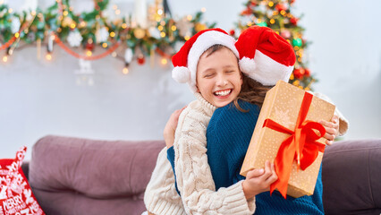 Fototapeta na wymiar happy and loving mother and son, dressed in a Santa Claus hat, hug at home, sitting on the sofa against the background of a Christmas tree. child and woman are laughing happily while holding gift box.