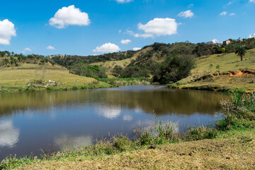 Fototapeta na wymiar Paisagem de verão em fazenda, com muita vegetação ao redor, lindo lago, céu azul tudo em meio a montanhas, no bairro Jardim das Oliveiras, Esmeraldas, Minas Gerais, Brasil.