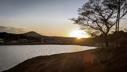P&ocirc;r do sol entre a silueta de uma &aacute;rvore ao entardecer com c&eacute;u dourado e  limpo, com reflexo em lago a no bairro Jardim das Oliveiras, Esmeraldas, Minas Gerais, Brasil