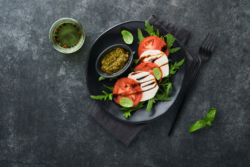 Caprese salad. Italian caprese salad with sliced tomatoes, mozzarella cheese, arugula, basil, olive oil in black plate over old brick tiles black background. Delicious Italian food. Top view.