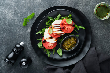 Caprese salad. Italian caprese salad with sliced tomatoes, mozzarella cheese, arugula, basil, olive oil in black plate over old brick tiles black background. Delicious Italian food. Top view.