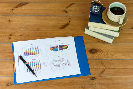 Business report,graph, put on wooden table,cop of coffee,alarm clock and books overlay on right side table. - Powered by Adobe