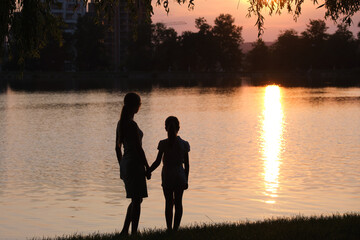 Happy mother and daughter standing together holding hands enjoying time in summer park in evening. Family love and relationship concept