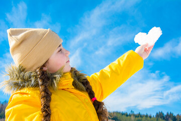 On a bright sunny day in winter, a little girl in a yellow jacket holds a heart made of snow in her hands against the blue sky.