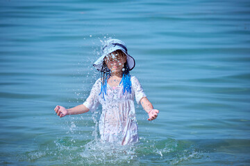 Little child girl in wet white dress and big hat playing alone in sea water enjoying summer...