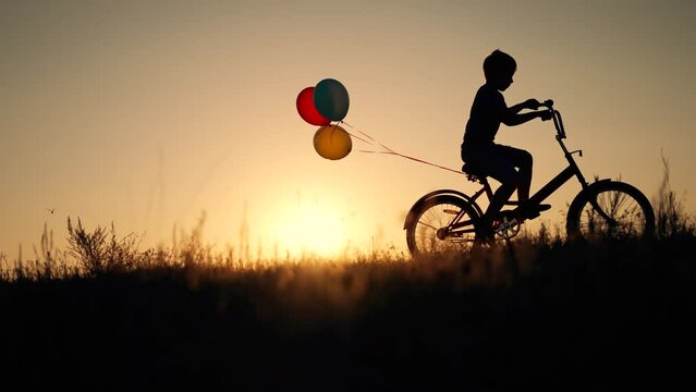 Dream child. Silhouette of kid on bike in park. Boy rides in park on green grass. Child games in nature.Traveling with balloons on bike.Active child freedom in summer.Boy learn to ride bike in nature.