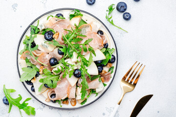 Tasty arugula salad with sweet pears, blueberries, roquefort cheese, smoked pork ham and crunchy walnuts. White kitchen table background, top view, space for text