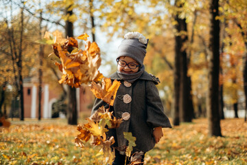 Happy cute brown-haired girl with down syndrome in fashionable coat and stylish eyeglasses tossing bright foliage up to the sky and laughing, kid enjoying time in warm autumn park, happy childhood