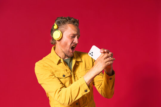 Singing handsome man enjoying his favorite song using phone and wireless headphones wearing jeans yellow jacket isolated on red background. Joyful man sing while recording his voice using smartphone
