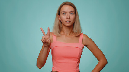 Blonde slim thin slender woman showing victory sign, hoping for success and win, doing peace gesture, smiling with kind optimistic expression. Young adult girl isolated on blue studio wall background