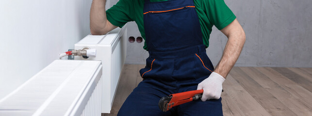 A male plumber installs a radiator in the heating system of an apartment. Guy in overalls and a gas...