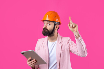 Portrait of young construction engineer wear orange hard hat, in a pink jacket standing on red studio background. A man with a tablet in his hands.