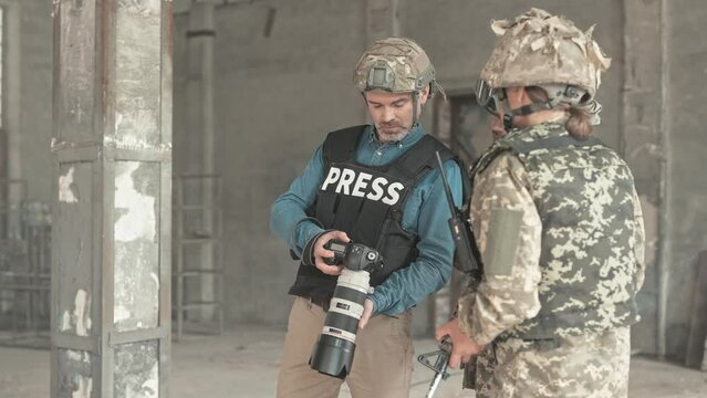 Close up of middle-aged professional photographer journalist working at war zone showing photos to soldiers in camouflage uniform with rifles in old ruined building hiding from bomb shelling