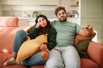 Bored couple of young people watching tv holding the remote control sitting on a sofa at home - Powered by Adobe
