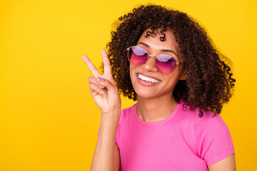 Photo of charming cheerful girl dressed pink t-shirt dark glasses showing v-sign empty space isolated yellow color background