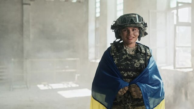 Portrait of Caucasian joyful beautiful female soldier in camouflage uniform covered in Ukrainian flag standing in ruined building. Pretty young brave woman paramedic smiling at camera. War in Ukraine