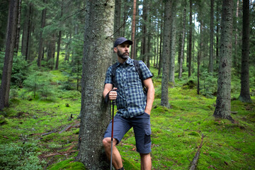 Hiker beard man in a green mountain forest. Dressed up in a cap, shorts, and plaid shirt with a backpack and trekking sticks