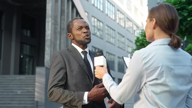 Rear of woman journalist with microphone stands outdoor interviewing African American successful businessman in suit. Male politician speaking with female reporter standing on street. Public relations