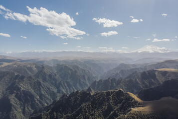 Panoramic view from a quadrocopter to the Caucasus mountain range and Mount Elbrus, in the foreground hills with green forests, sunny September day with clouds