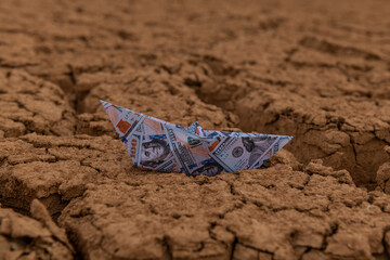 A paper boat with an image of a 100 American dollar bill stands on cracked dry clay in the desert