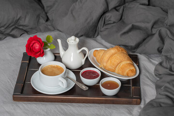 Breakfast in bed with espresso coffee, croissant, honey and jam on the wooden tray, close up. White cup of coffee on the gray bedsheet with pillow and blanket