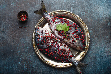 Two raw fishes dorado seasoned in pink peppercorn ready to be cooked laying on rustic metal plate top view. Healthy cooking, diet food eating concept. Dark blue concrete stone background