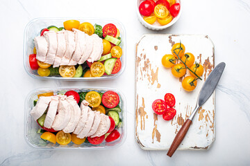 Healthy meal prep for lunch, diet boiled chicken fillet with vegetables salad in lunch boxes containers top view on white marble kitchen table background with knife and cutting board. Space for text