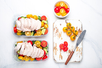 Healthy meal prep for lunch, diet boiled chicken fillet with vegetables salad in lunch boxes containers top view on white marble kitchen table background with knife and cutting board. Space for text
