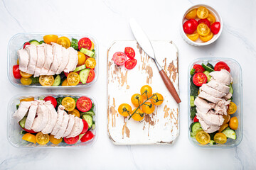 Healthy meal prep for lunch, diet boiled chicken fillet with vegetables salad in lunch boxes containers top view on white marble kitchen table background with knife and cutting board. Space for text