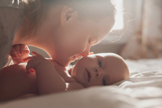 Mom Kisses And Hugs A Smiling Newborn Baby Lying On The Bed. Young Woman With A Child At Home