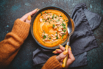 Female hands in yellow knitted sweater holding a bowl with pumpkin cream soup on dark stone...