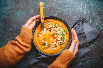Female hands in yellow knitted sweater holding a bowl with pumpkin cream soup on dark stone...