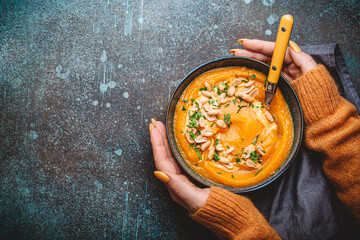 Female hands in yellow knitted sweater holding a bowl with pumpkin cream soup on dark stone...