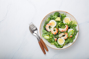 Healthy keto-friendly salad high in protein and healthy fats with shrimps, avocado and boiled eggs in ceramic boil on white marble table background from above, diet and clean eating concept