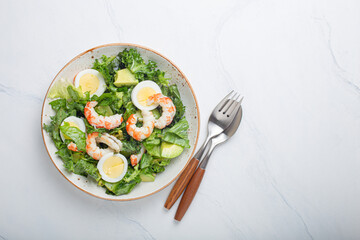 Healthy keto-friendly salad high in protein and healthy fats with shrimps, avocado and boiled eggs in ceramic boil on white marble table background from above, diet and clean eating concept