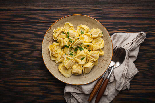 Italian filled tortellini ravioli on ceramic plate top view on dark rustic brown wooden table background with fork and spoon. Traditional pasta dish of Italy, space for text