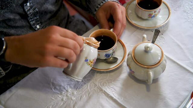 Close up of interracial young couple's hands preparing and drinking a coffee with milk and sugar using a vintage porcelain cup with flowers pattern.
