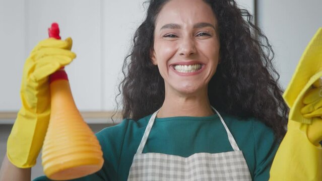 Happy cleanup worker in yellow rubber gloves holding detergent and sponge for wiping dust. Housewife playful splashing pure liquid from bottle at camera enjoying housekeeping chores everyday cleanness