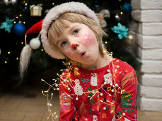 child in Christmas pajamas, Santa hat and a deer make-up painted on his face makes funny faces....