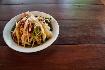 Close-up to papaya salad in a small plate on a wooden table placed on the left side of the picture.