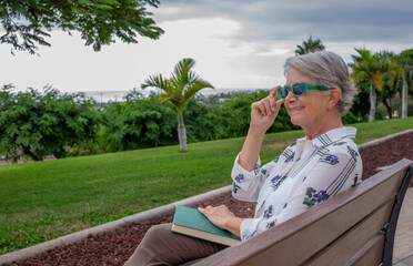 Portrait of beautiful senior woman with green sunglasses sitting on a bench in public park with a book over her legs - caucasian lady enjoying free time and retirement